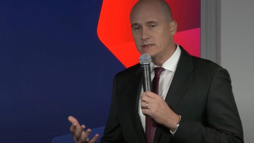 Mirek Dusek in a suit speaking into a microphone with a red and blue background.