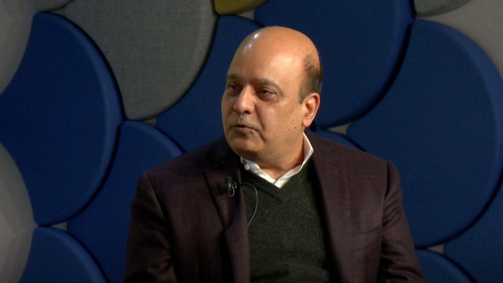 Pervinder Johar speaking, wearing a dark blazer and sweater, with a microphone attached, against a geometric patterned background.