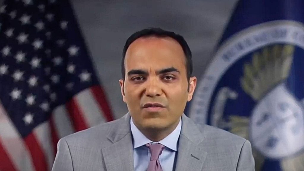 Rohit Chopra in a suit speaking, with the U.S. flag and a blue emblem in the background.