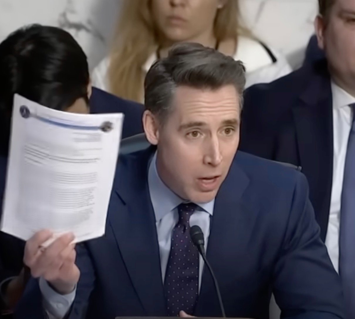 Josh Hawley in a suit holds up a document during a speaking engagement, with other people in the background.