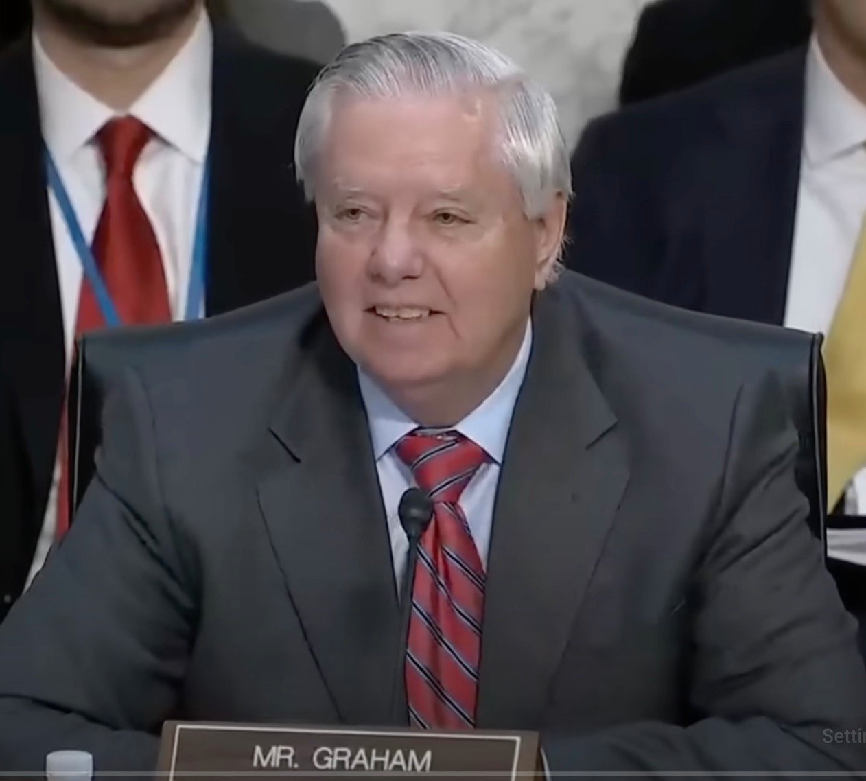 Lindsey Graham sitting at a desk speaking into a microphone, wearing a suit and red striped tie, with a nameplate labeled "Mr. Graham."