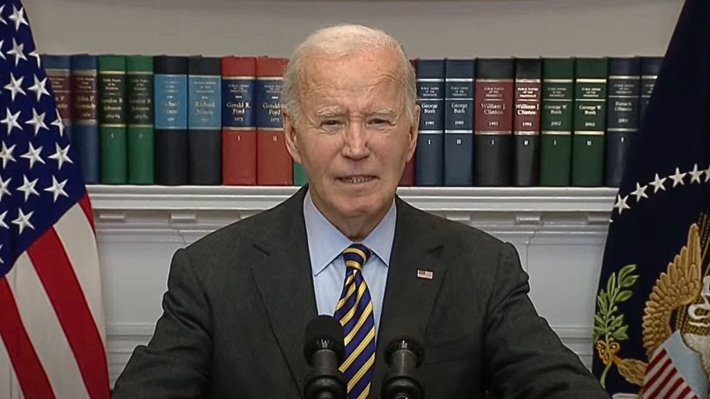 Biden in a suit and striped tie speaks at a podium in front of bookshelves, an American flag, and a seal with a bald eagle.