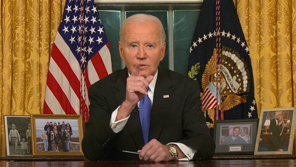 Biden in a suit giving a speech in a formal setting with U.S. flags and framed photographs in the background.