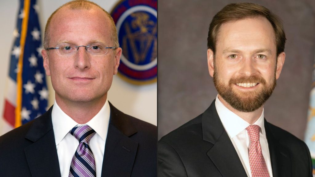 Carr and Ferguson in formal suits with ties, both smiling. The man on the left is bald with glasses, and the man on the right has a beard. An American flag is partially visible in the background.