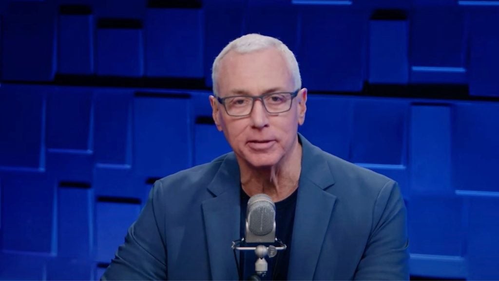 Dr. Drew with glasses speaking into a microphone, blue background.