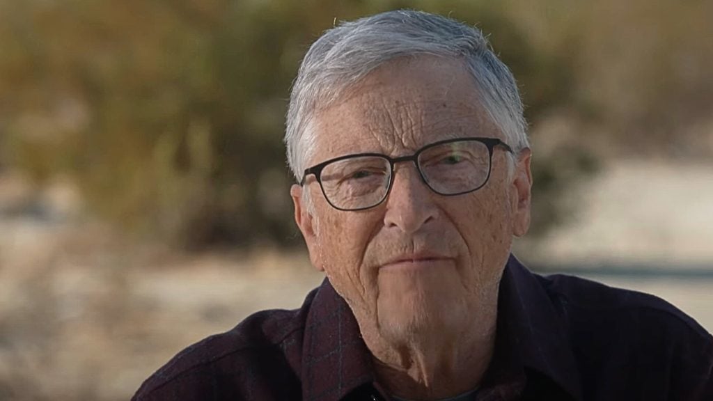 Bill Gates with gray hair and glasses wearing a dark shirt, standing outdoors.