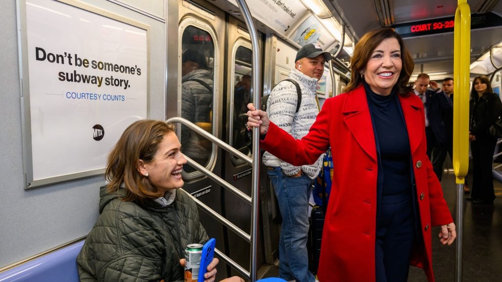 Hochul in a red coat stands smiling in a subway car next to a seated passenger, with a sign on the wall reading "Don't be someone's subway story. Courtesy counts."