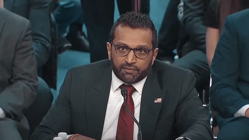 Kash Patel wearing glasses, a suit with a red tie, and speaking into a microphone at a formal event.