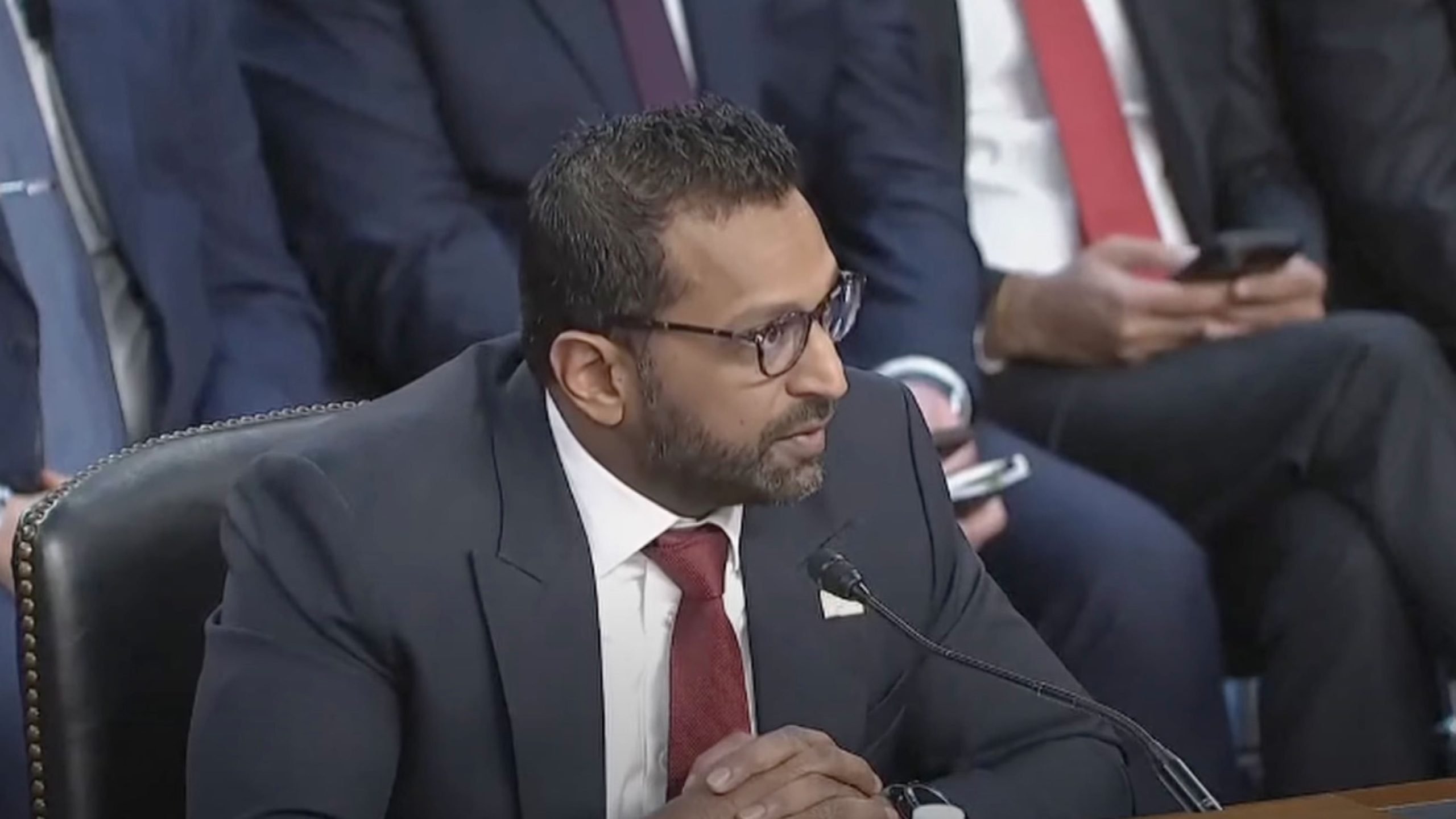 Kash Patel in a suit and red tie speaking into a microphone, seated at a table during a formal event, with others in suits in the background.