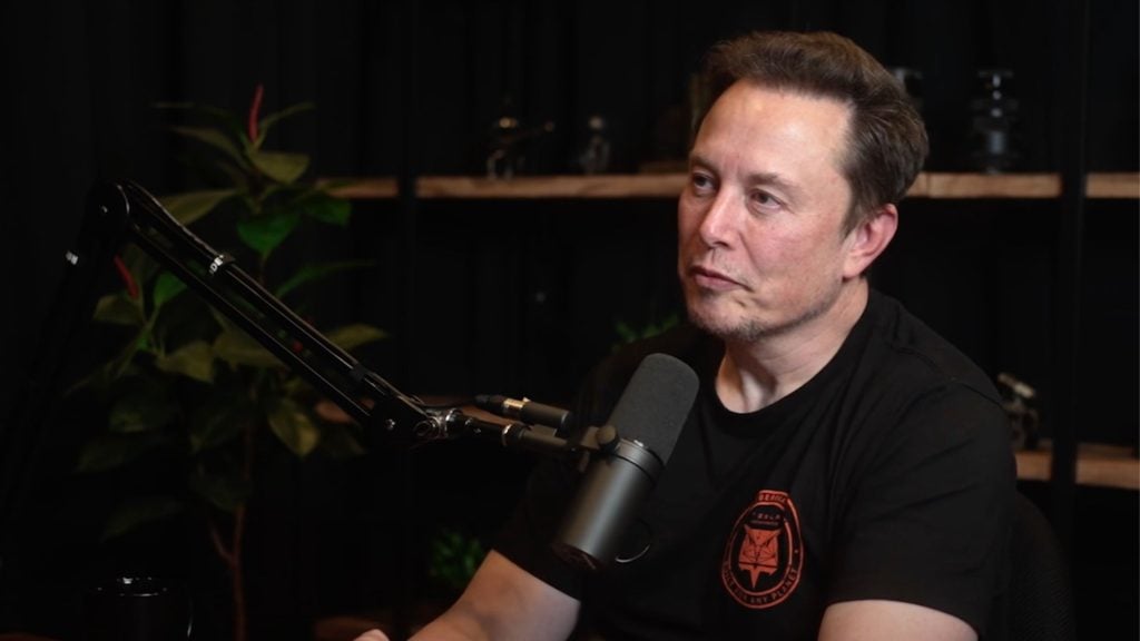 Elon Musk sitting in front of a microphone, wearing a black t-shirt with a logo, in a dimly lit room with plants and shelves in the background.