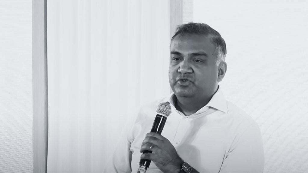 Mohan holding a microphone, speaking in front of a white background.