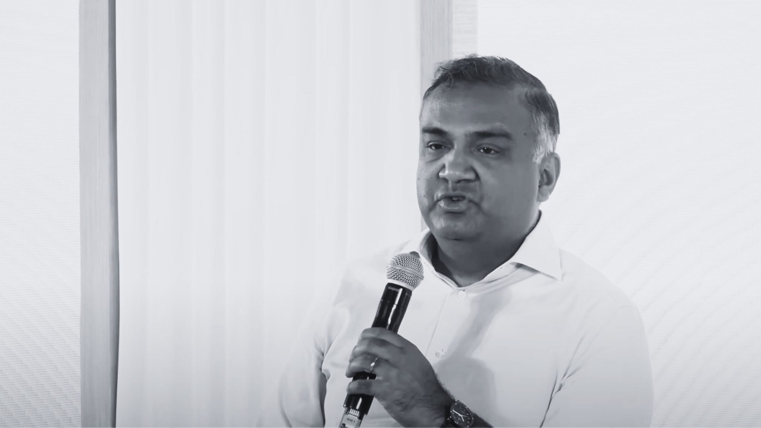 Mohan holding a microphone, speaking in front of a white background.