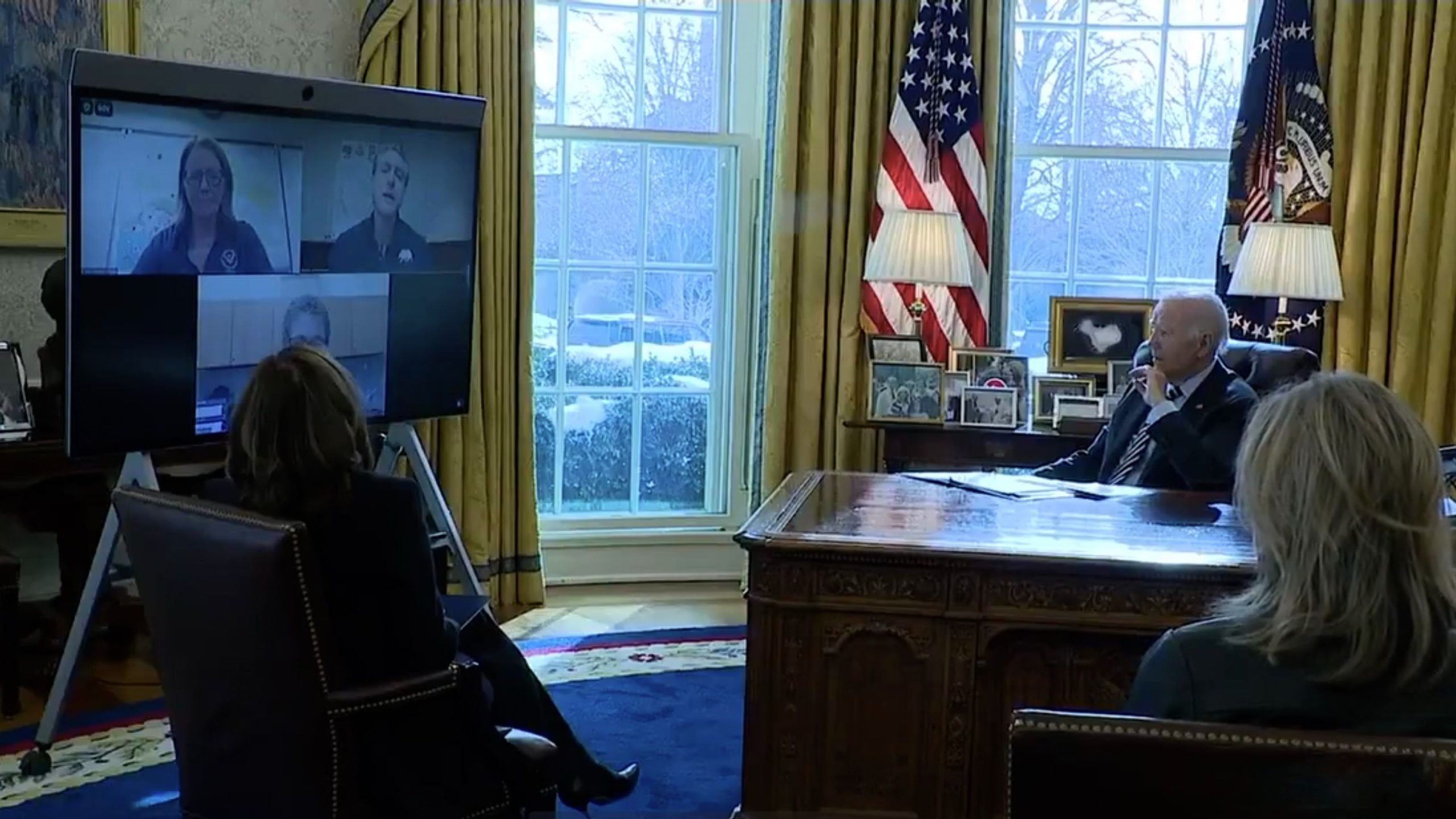 Biden sitting in an office watching a video conference on a large screen, with several people displayed on it.