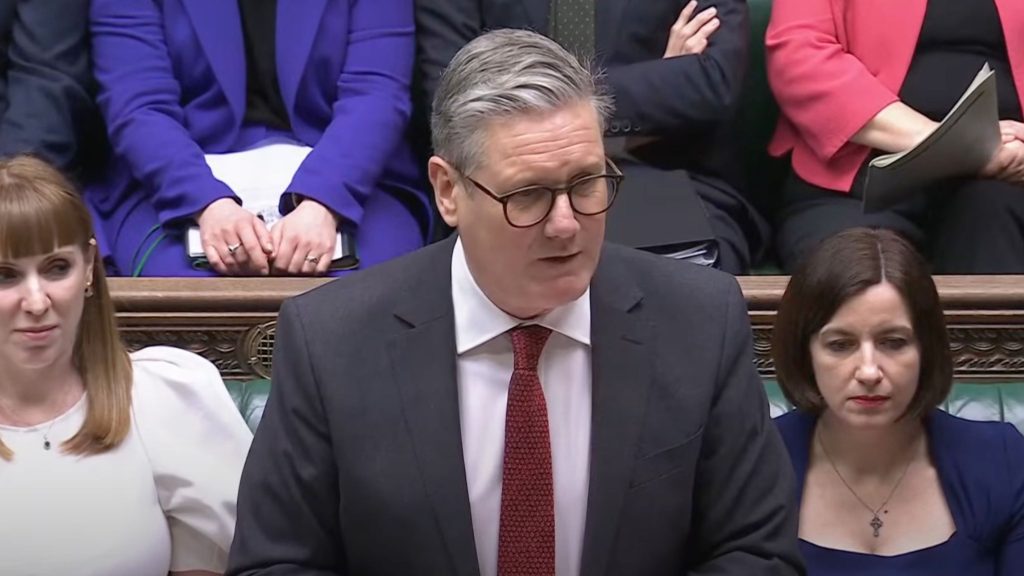 Starmer with glasses and a red tie speaks at a government meeting, flanked by people in formal attire.