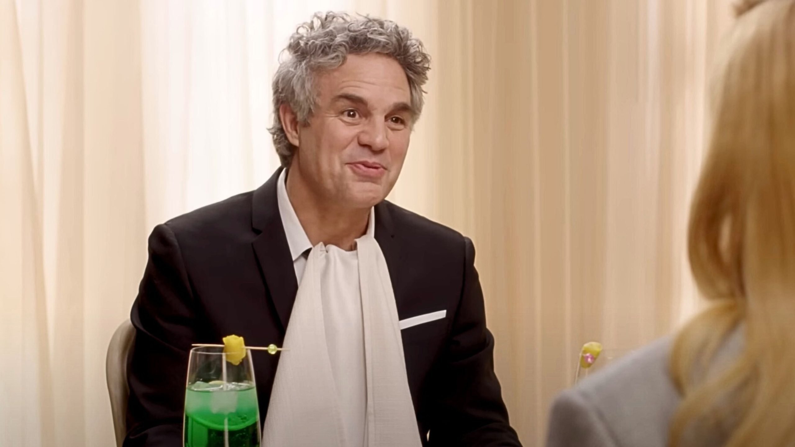 Mark Ruffalo in a suit with a white scarf sits at a table with a green cocktail garnished with a lemon slice.