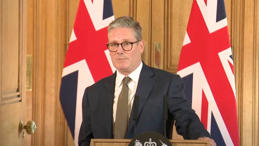 Starmer in a suit and glasses speaking at a podium with UK flags in the background.