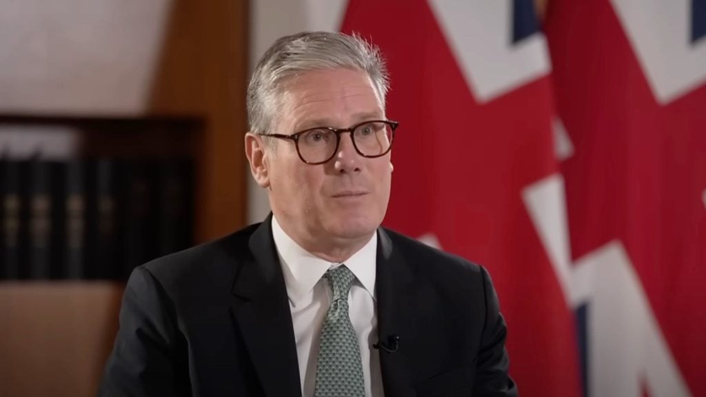 Starmer in a suit and tie, wearing glasses, speaking in front of a blurred background with partial red and white flag design.