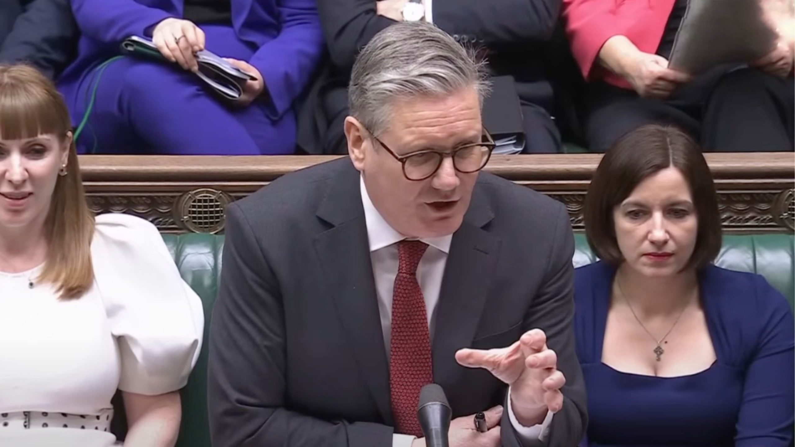 Starmer in a suit is speaking into a microphone, seated between two women in a formal setting.