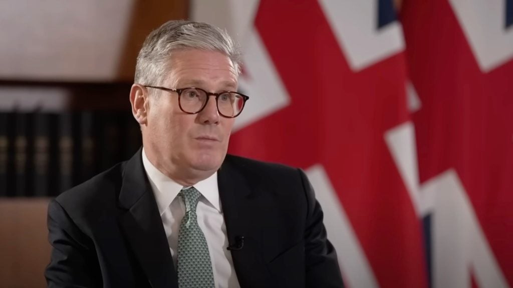 Starmer in a suit and glasses speaking, with the UK flag in the background.