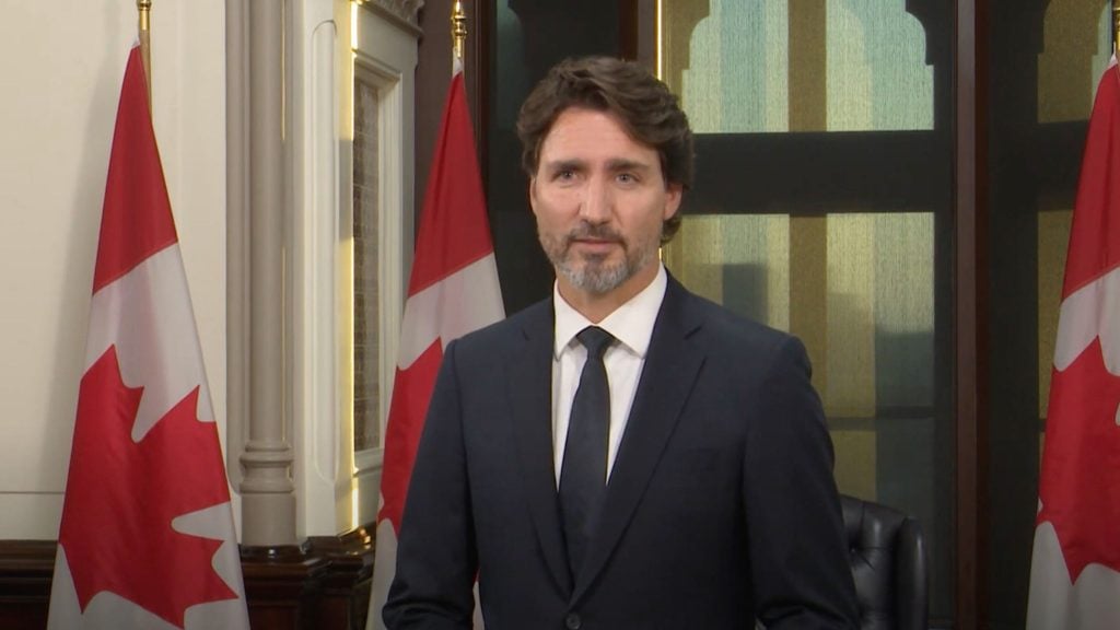 Trudeau in a suit stands between two Canadian flags in an indoor setting.