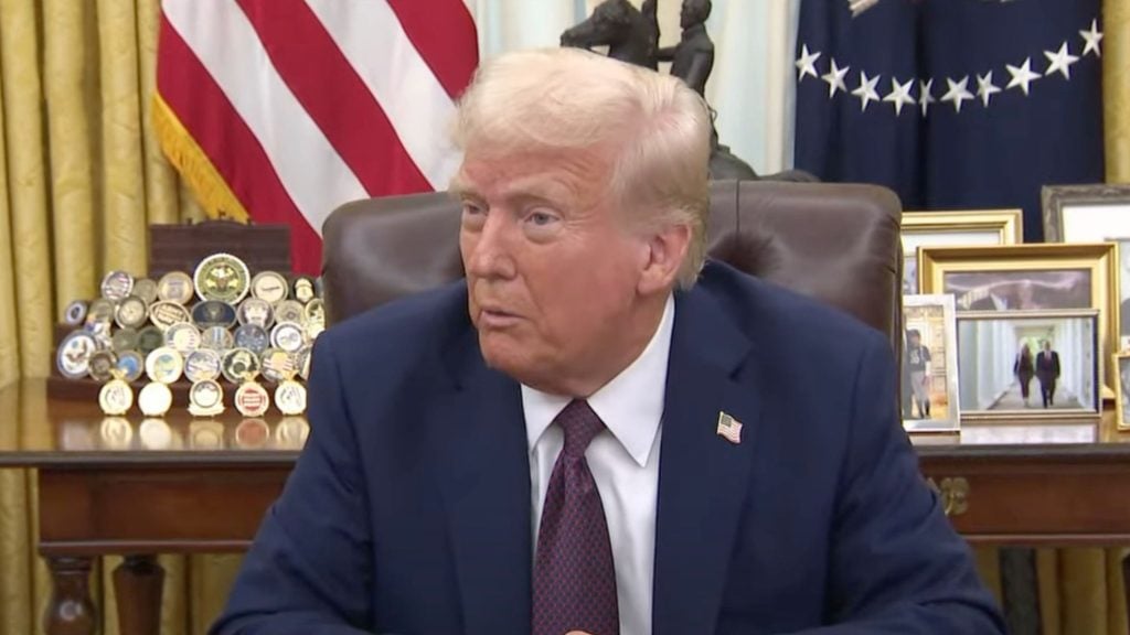 Trump seated at a desk with a background of a U.S. flag, framed pictures, and challenge coins.