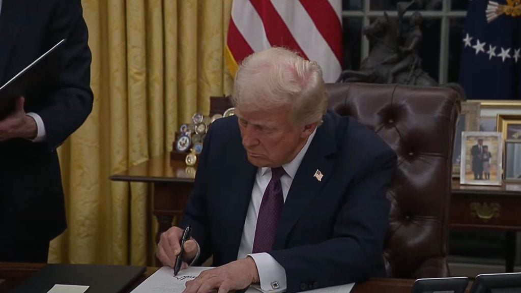 Trump seated at a desk in an office with yellow curtains, writing with a pen on a document.