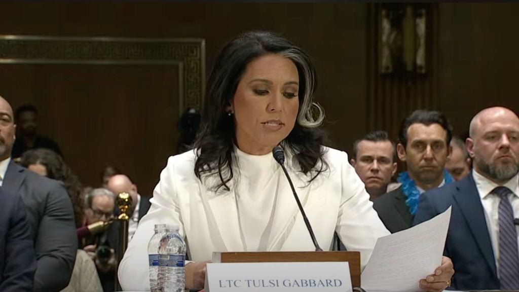 LTC Tulsi Gabbard in a white suit speaks into a microphone with a paper in hand, seated at a table with a nameplate and a bottle of water.