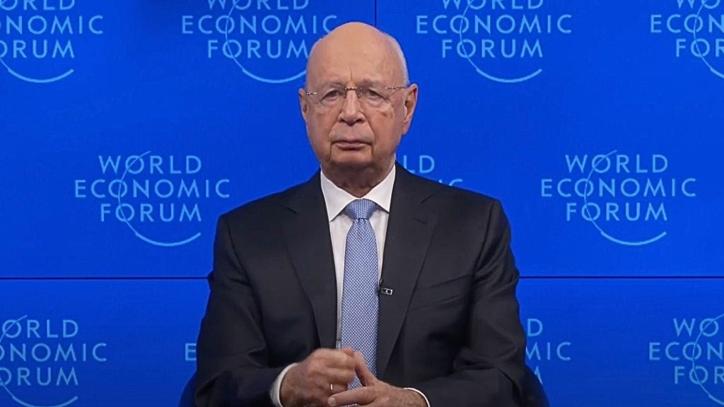 Schwab in a formal suit and tie speaking at the World Economic Forum, with a blue background displaying the forum's logo.