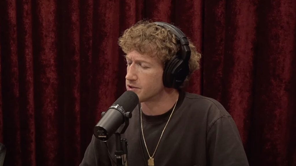 Zuckerberg speaking into a microphone with headphones on, in front of a red curtain.