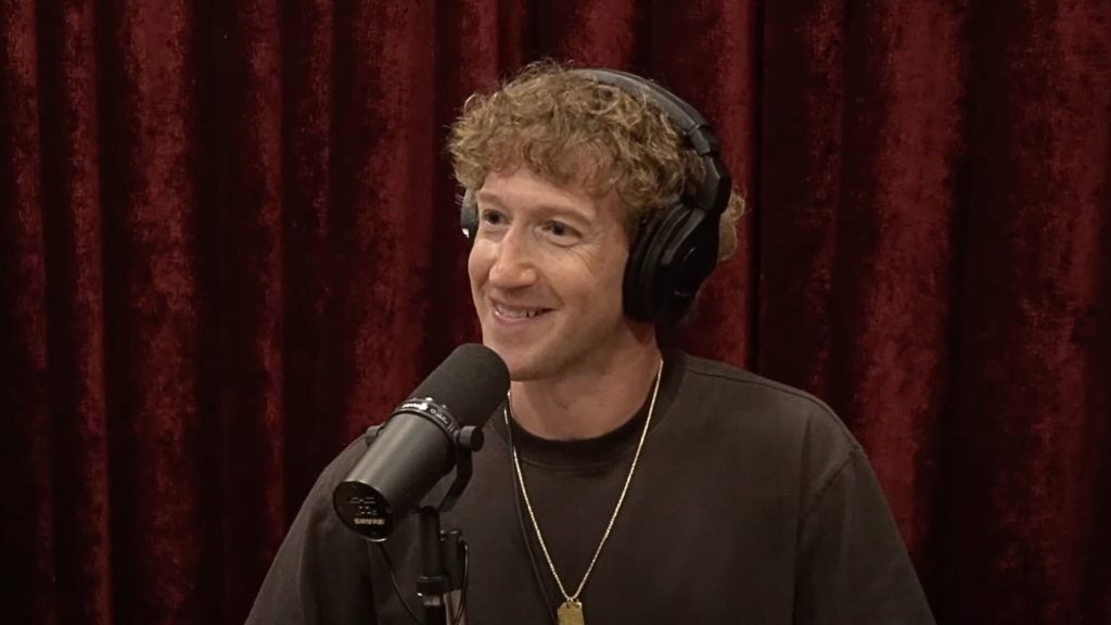 Zuckerberg with curly hair wearing headphones and speaking into a microphone against a red curtain backdrop.