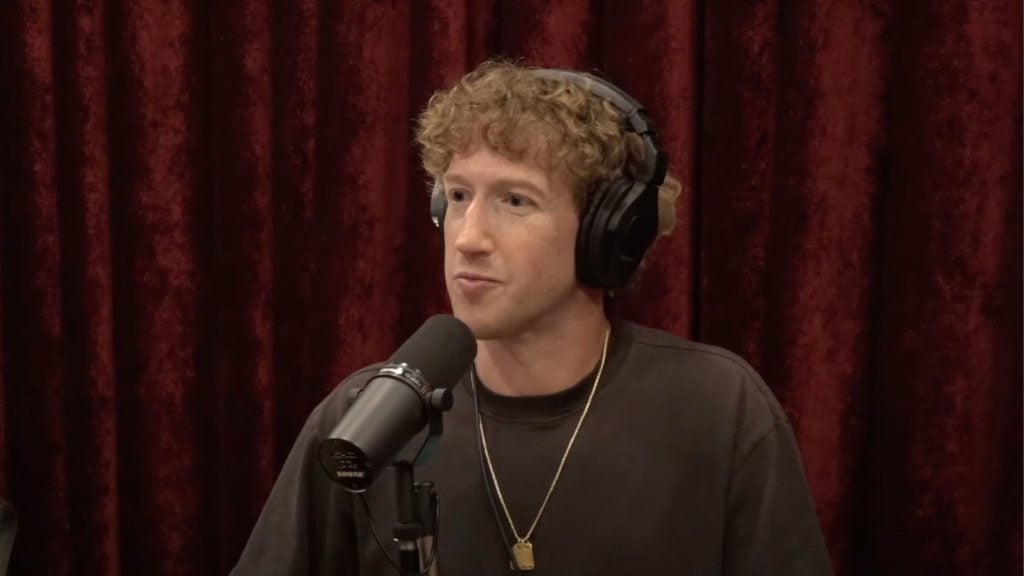 Zuckerberg with curly hair wearing headphones and speaking into a microphone, in front of a red curtain backdrop.