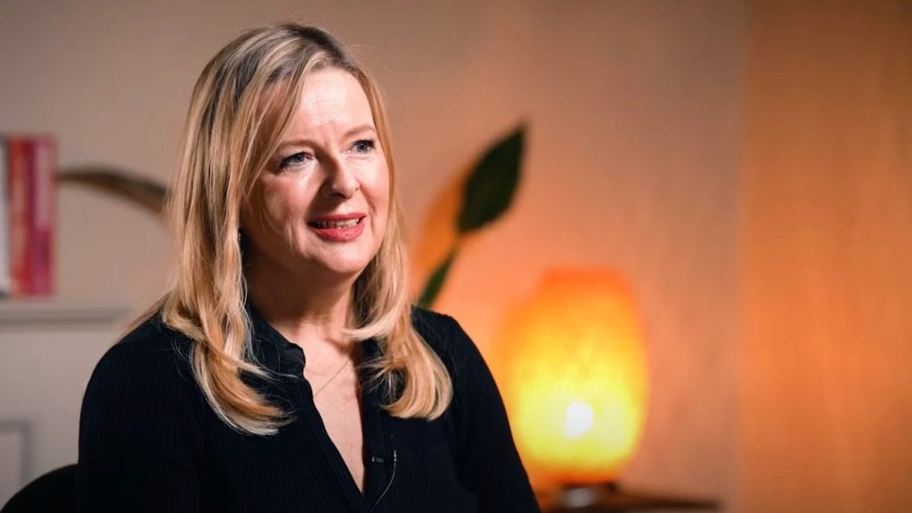 Pearson with long blonde hair is smiling softly, seated indoors with a warm-colored lamp in the background.