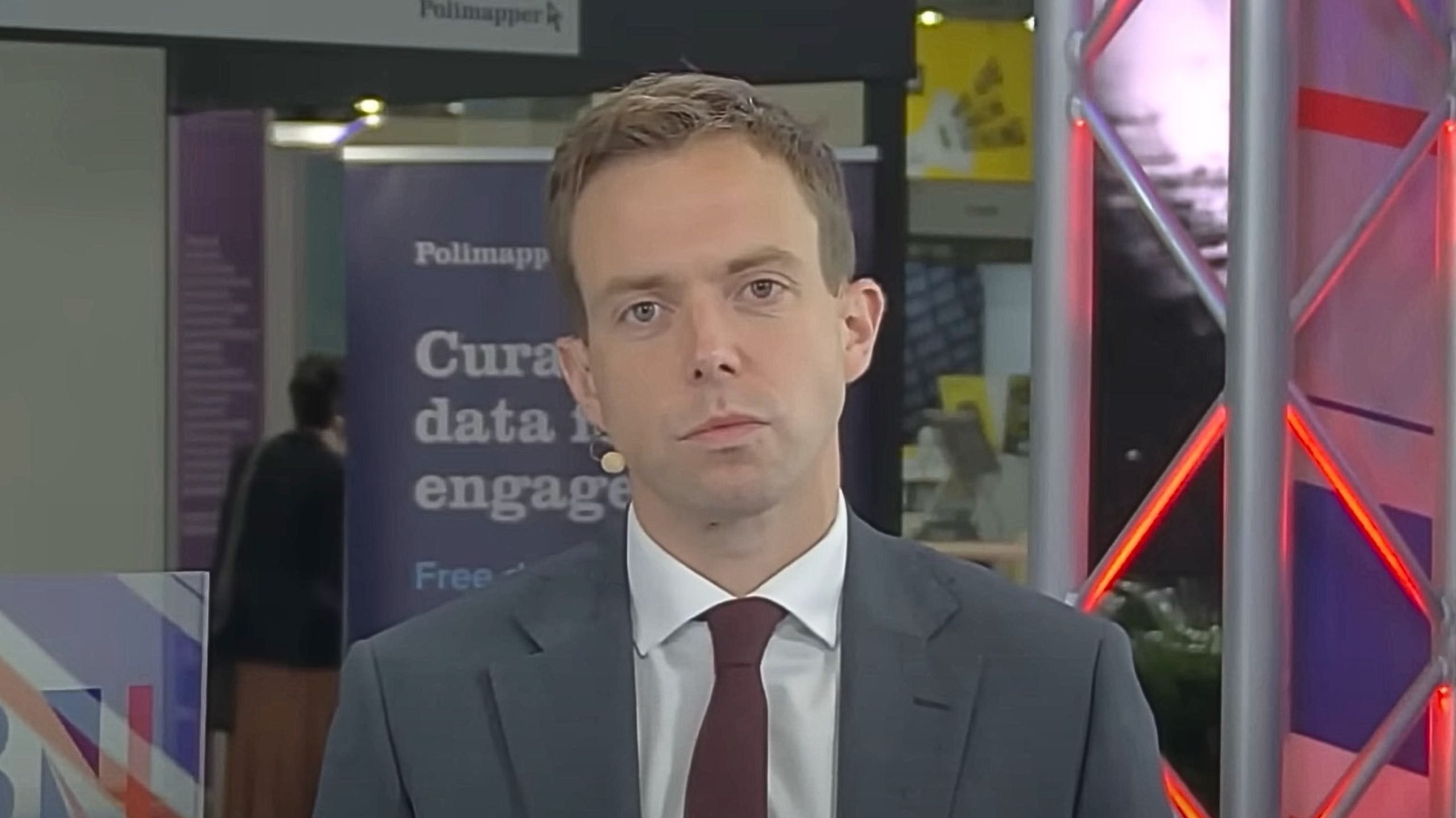 Jake Richards in a suit and tie standing in front of a backdrop that includes text and signs, with a microphone visible on his cheek.