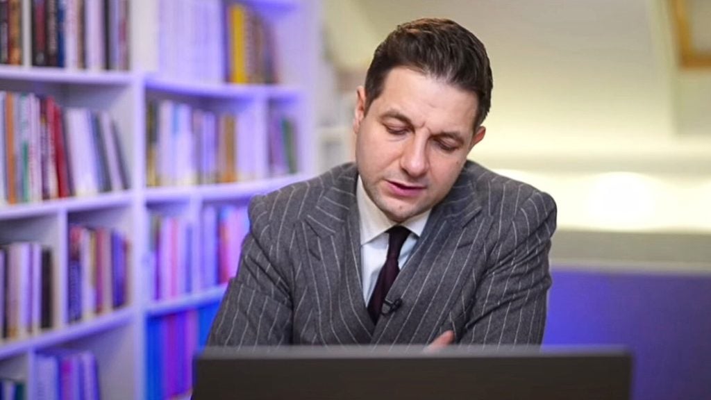 MEP Patryk Jaki in a gray pinstripe suit sitting at a desk, looking down at a laptop, with bookshelves in the background.