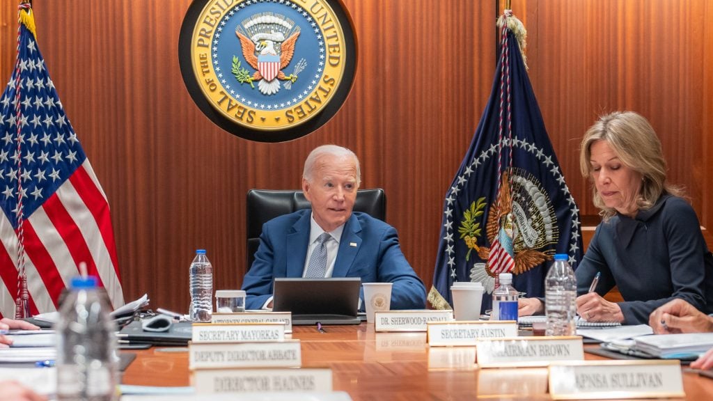 Biden in a blue suit and a woman writing notes sit at a conference table with official nameplates and a laptop, surrounded by American flags and a presidential seal on the wood-paneled wall.