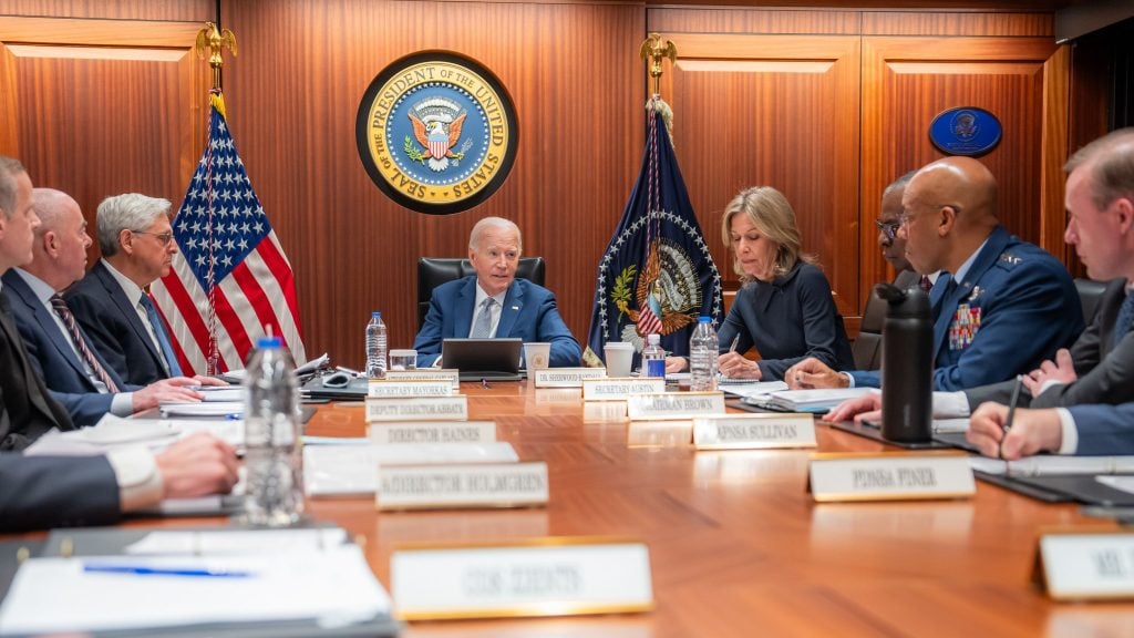 Biden and officials seated at a conference table in a formal meeting room, with the Presidential seal and American flags in the background.