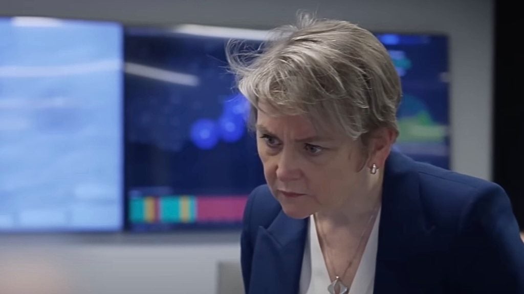 Cooper with short hair wearing a suit looks intently at something off-camera, in a room with screens displaying data in the background.