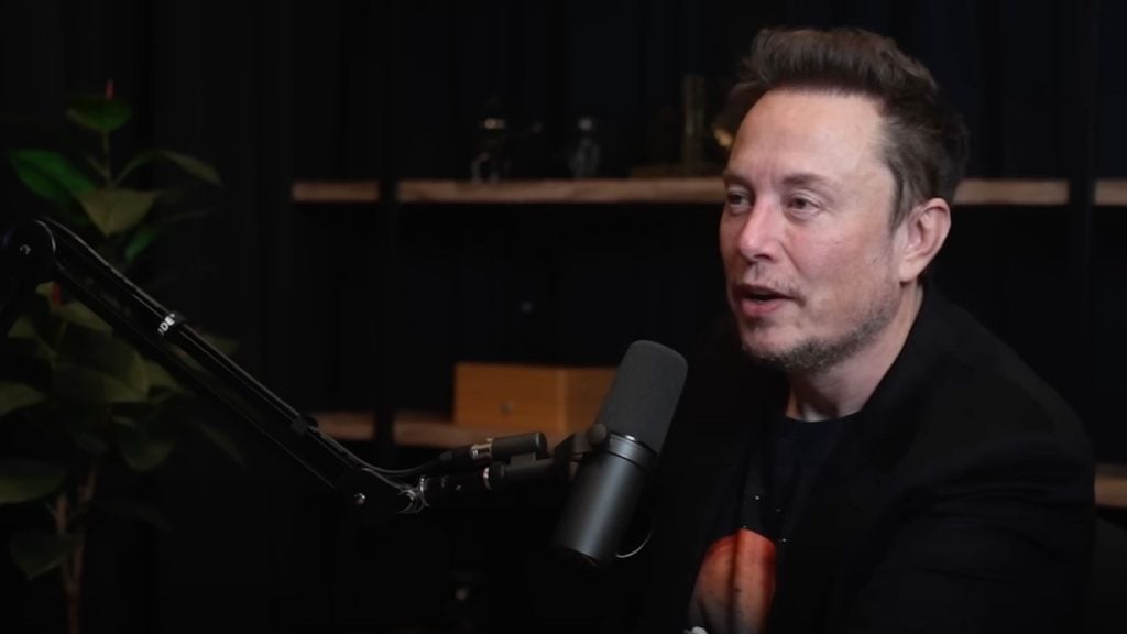 Elon Musk speaking into a microphone with a dark background and a plant.