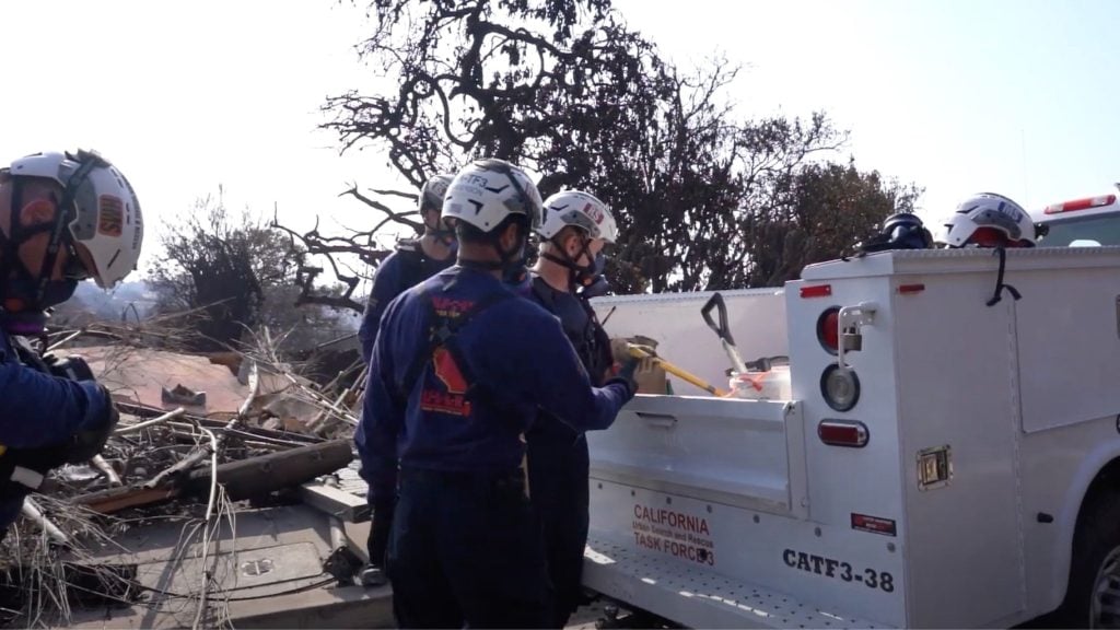 Rescue workers wearing helmets and protective gear stand by a utility truck with debris and fallen branches surrounding them, as part of a search and rescue operation by California Task Force 3.