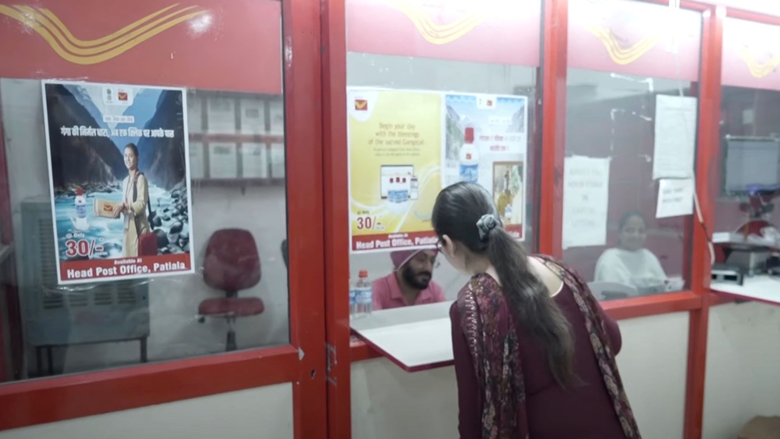 A woman at a post office counter interacting with an employee. Posters promoting postal services are displayed on the glass window divider. The setting appears to be the Head Post Office in Patiala.