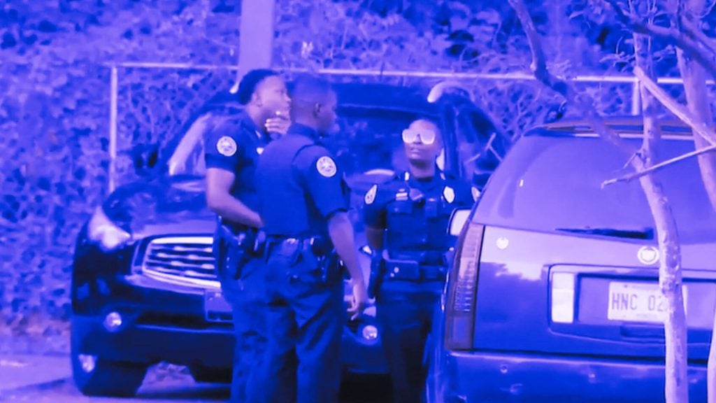 Four police officers in uniform stand near a dark vehicle, engaged in conversation, in an outdoor setting with trees and foliage in the background.