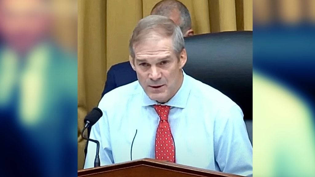 Jim Jordan in a white shirt and red tie speaking into a microphone at a podium in a formal setting with a blurred background.