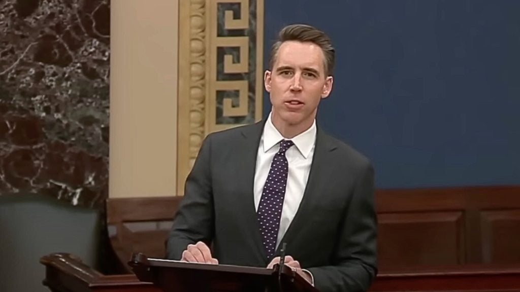 Josh Hawley in a suit speaking at a podium inside a formal setting, such as a legislative chamber.