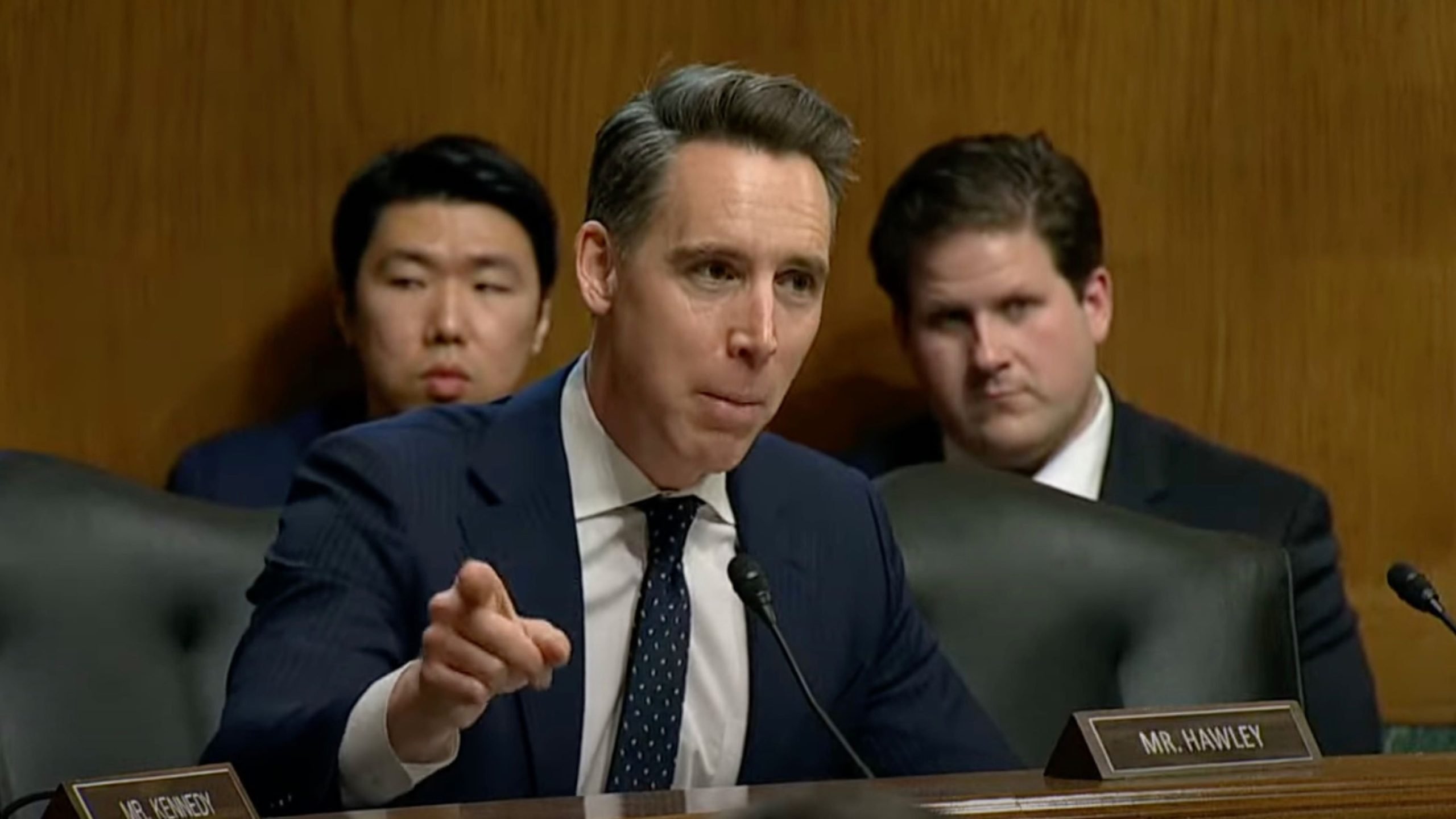 Josh Hawley in a suit speaking into a microphone during a formal meeting, with other individuals seated behind him.