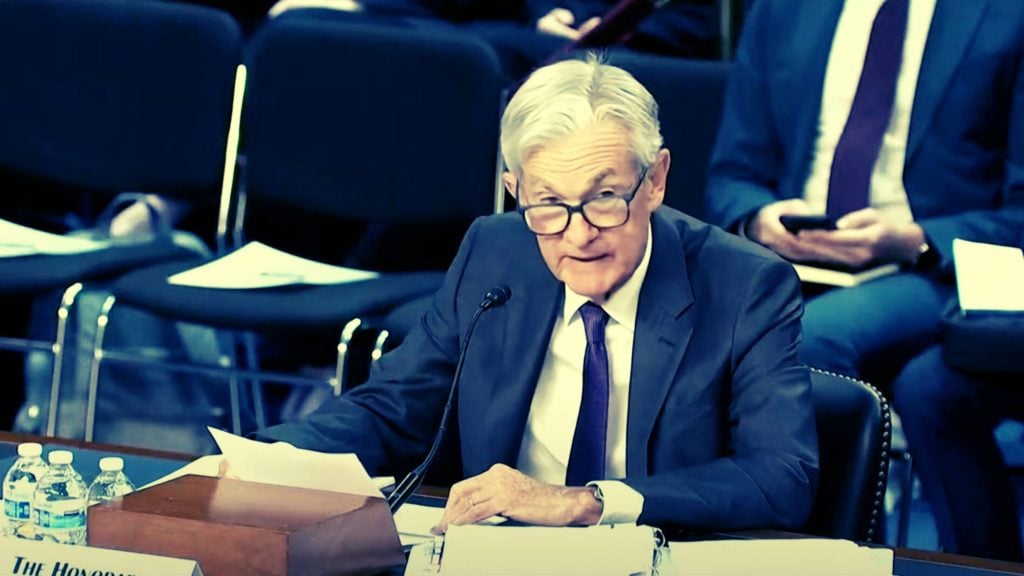 Jerome Powell in a suit and glasses speaking into a microphone at a conference or hearing, with papers and water bottles on the table.