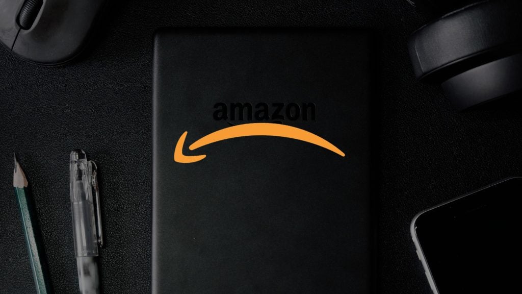 A black notebook with the Amazon logo, featuring the word "amazon" and a curved arrow, placed on a dark surface alongside a pen, a pencil, and part of a smartphone.