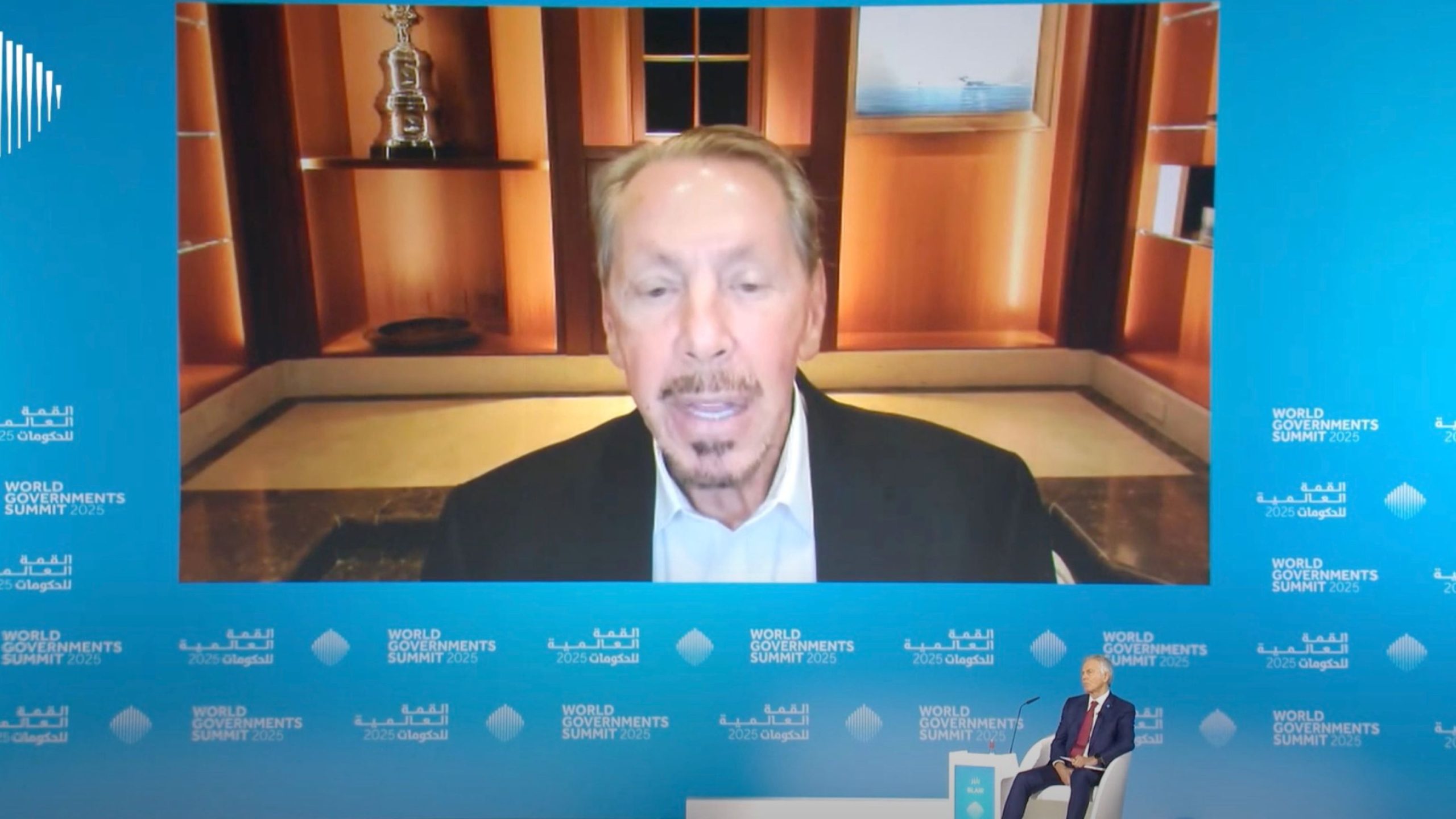 A large screen showing Larry Ellison speaking at the World Government Summit 2025, with a seated Tony Blair in the foreground.