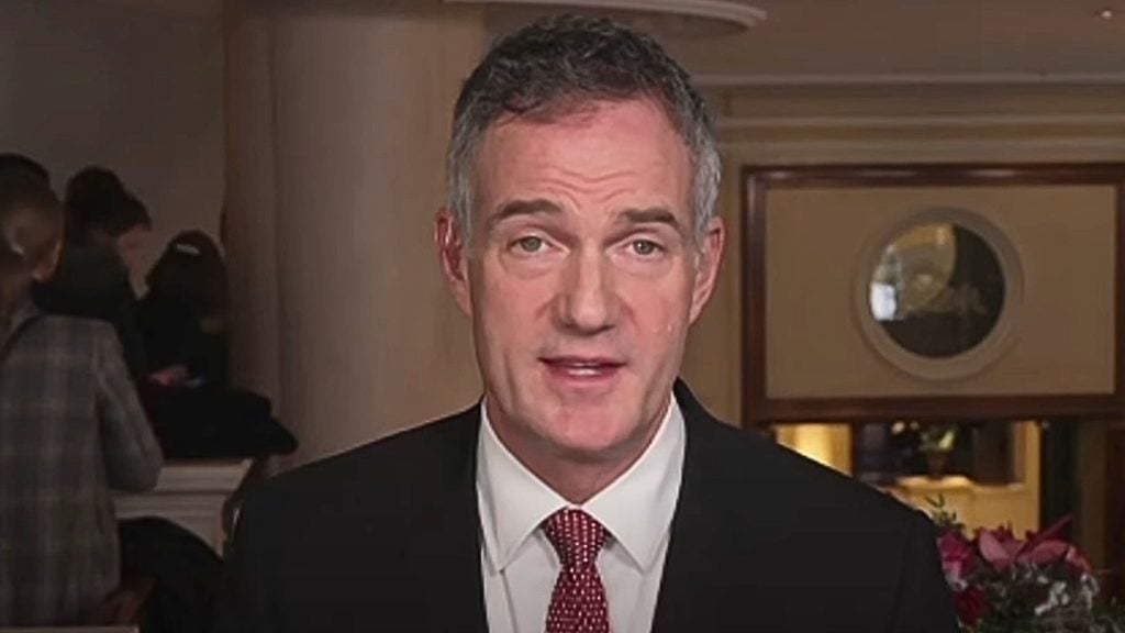 Peter Kyle in a suit and red tie speaking indoors with a serious expression; behind him, there is a blurred background of people and a decorative mirror.