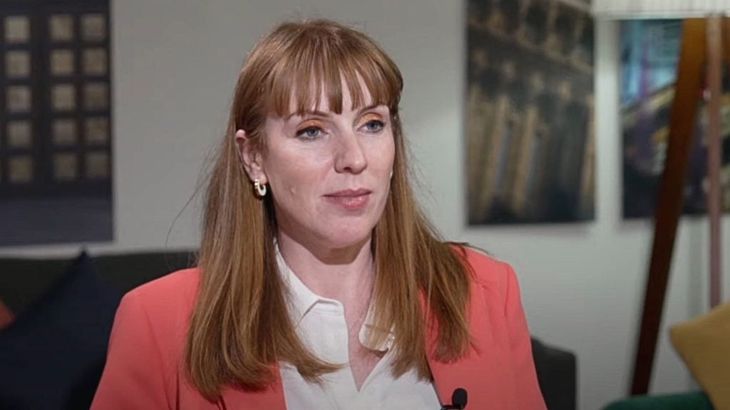 Rayner with long auburn hair wearing a salmon-colored blazer and a white shirt, sitting indoors with framed pictures on the wall behind.