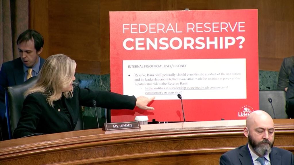 Senator Cynthia Lummis (R-WY) sits at a desk with a microphone, next to a placard labeled "Ms. Lummis" while pointing at a large document in the background which contains a note surrounded by a red border. The document shows a quote from a confidential Federal Reserve implementation handbook on account actions that requires Federal Reserve staff to "consider the conduct of the institution and its leadership and whether association with the institution poses reputational risk to the Reserve Bank."